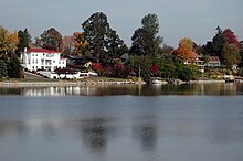 Houses with neat lawns, gardens, and, in some cases, boat docks, face a lake. A white, three-story house with a red roof stands out because of its size and color and because the other houses are mostly hidden by trees and shrubbery. Some of the foliage is red or golden, the rest green. The lake is calm, the sky clear.
