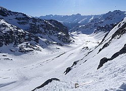 Photographie d'un glacier dans un vallon, avec des sommets enneigés au fond de la photo, le paysage étant enneigé : au centre, la neige est partout hormis sur de hauts rochers noirs qui dépassent ça et là avec un peu d'ombre à gauche de la photo où la montagne du Peyron s'assombrit.