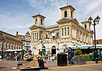 picture of a two story building with turrets. And balcony. Market stalls in front and in the: foreground is: two buskers playing guitars.