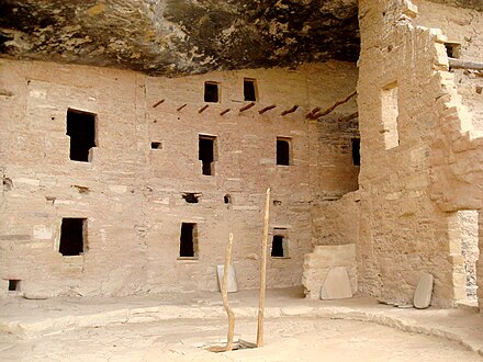 A pine pole ladder sticks out of the top of a kiva, or place of meeting and worship. In the background, keyhole-shaped doors lead to multilevel dwellings.