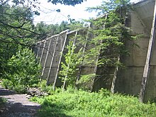 Foto einer Betonmauer mit Strebepfeilern, die durch ein Waldgebiet mit einem Pfad nach links führt.  Tief liegende Vegetation befindet sich am Fuß der Mauer.