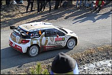 Fugleperspektiv av Sébastien Loebs Citroën DS3 WRC, hvit, rød og gullfarget, på en asfaltert vei omgitt av tilskuere.