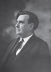 A portly man with wavy, black hair and a prominent nose, wearing a black jacket and tie and white shirt