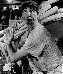 A man in a light baseball uniform and dark cap holding several baseball bats as if ready to swing