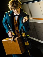 Jeune homme blond aux cheveux courts vu de face, portant un manteau de couleur bleu canard, une écharpe jaune et marron, une valise en cuir dans sa main droite et une baguette magique tendue dans sa main gauche. Il regarde le photographe.