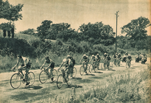 Photo en noir et blanc d'un peloton cycliste sur une route de campagne bordée de champs.