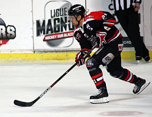 Photographie d’un homme, de tronc, portant un équipement de hockey sur glace, un maillot à dominante noire, le regard un peu dans le vide