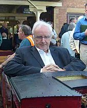 Une photographie de Pete Waterman, un homme âgé de race blanche aux cheveux blancs, vêtu d'un costume, assis les bras croisés