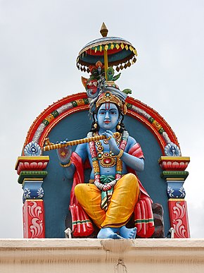 Statua di Khrisna nel tempio di Sri Mariamman a Singapore, spesso raffigurato come suonatore di flauto. Ha una corona regale (kirīṭa mukuṭa) con penne di pavone (mayūrapattra) che simboleggiano l'immortalità, in quanto il progenitore di questo animale nacque da una piuma di Garuḍa. La ghirlanda di Krishna è una ghirlanda di fiori (tulasī) ed è composta da cinque filari di fiori che rappresentano i cinque sensi dell'uomo. La sua postura è la ardhasamasthānaka pādasvastika, la postura a gambe incrociate con il piede destro che tocca con le punte delle dita il terreno mostrando leggerezza e calma.