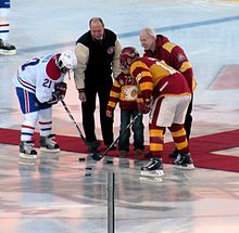 Two hockey players lean forward, facing each other, as three people standing on a carpet have each just dropped a puck to the ice.