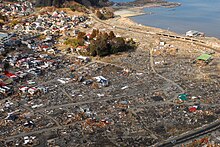 Photo couleur : vue aérienne d'une zone habitée de bord de mer, dévastée après un séisme et le passage d'un tsunami.