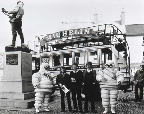 c. 1965–1970, view of old fashioned Michelin omnibus and two michelin men with bystanders behind Charles Rolls statue, Monmouth, Wales.