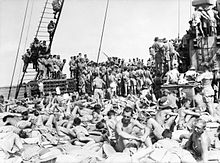 Black and white photo of a large number of men crowded together on the deck of a ship. Those in the foreground are sunbaking with their shirts off. Those in the background are standing with their backs to the camera. Some of the men in the background have climbed the ship's rigging or onto structures on the deck.