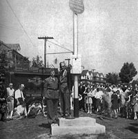 A man in a suit standing on a stage next to a large pole. There is a Marine in the background in his dress uniform and behind him a crowd of people are watching the man on the stage.