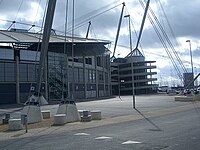 A grey stadium exterior with glass fronting. Adjoining it is a spiral walkway made of concrete, rising almost to the full height of the structure.