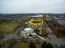 Overhead view of the New York State Pavilion on a cloudy day in 2017. The Theaterama is visible in the left foreground, while the observation towers are in the right foreground. Behind them is the Tent of Tomorrow, the top of which is painted yellow. Roads are visible on all sides.
