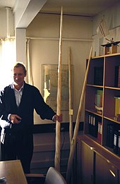 Un homme en costume se tient debout à gauche de la photo, tenant une longue dent de narval torsadée de la main droite. L'objet dépasse très largement la taille de l'homme et atteint presque le plafond.