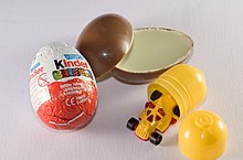 Thin egg-shaped chocolate shell, broken open to reveal a large plastic capsule that contains a cheap plastic toy