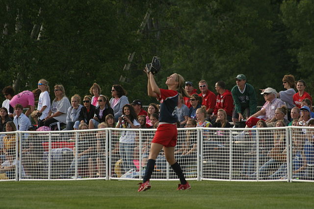 A light-skinned woman, wearing a red and blue shirt and red shorts, has her arms in the air to catch a ball in a grassy field as spectators look on.