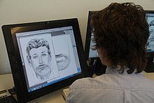 Photo couleur d'une personne, vue de dos, assise devant un écran d'ordinateur dans lequel un logiciel de dessin est ouvert et affiche le portrait noir et blanc d'un homme barbu.