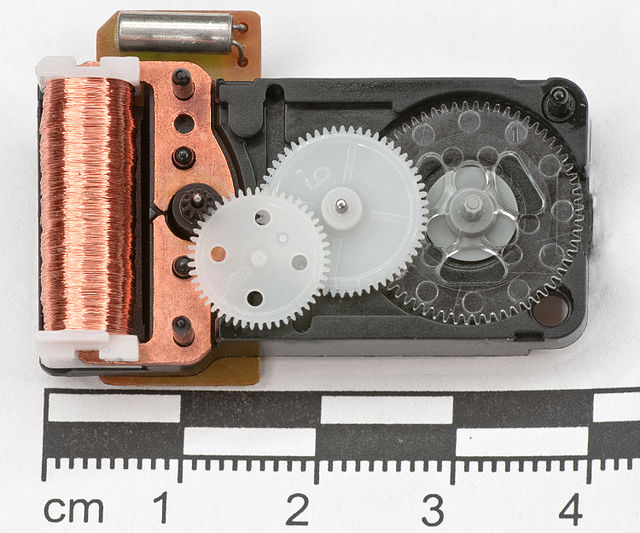 Disassembled analog quartz clockwork; quartz crystal oscillator (top left), Lavet-type stepping motor (left) with a black rotor sprocket and connected