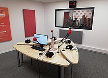 une salle d'enregistrement avec 3 tables et 5 microphones, un ordinateur et au fond derrière la vitre la régie, aux murs des formes décorées permettant d'abattre les sons parasites