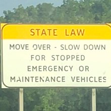 A large white sign bears a bright yellow header with all capital lettering that's slight peeling indicates wear by the elements and old age. The words "State Law" fill the yellow header in bold black print typical of road signs. Below it is written, slightly smaller, but still very large "Move over - slow down for stopped emergency or maintenance vehicles." The sign is held up by two large posts driven into the ground (not visible). In the background is a blue sky and green foliage from trees and bushes that line the highway.