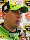 A man in his thirties being interviewed by the media. He has a green and black baseball cap on his head and is wearing identically colored racing overalls.