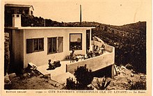 Postal en blanco y negro.  Vista a un edificio de una sola planta en la ladera con una terraza donde los adultos se sientan en las mesas y un niño en un coche de pedales.