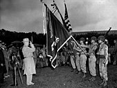 Man in an overcoat and fedora salutes a unit color's with streamers.