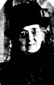 An older white woman, wearing a black hat and coat, and glasses