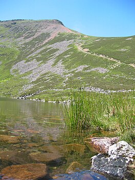 A clear lake in the foreground with a steep mountain ridge beyond