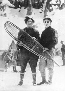 Frontale Schwarzweißfotografie von zwei lächelnden Männern, die gemeinsam Schneeschuhe aus Holz vor sich halten. Die Haare des linken Mannes stehen ab und der rechte Mann hat Wellen. Im Hintergrund sind einige Menschen, eine Hütte und eine Winterlandschaft.