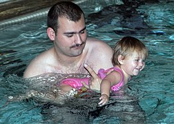Un homme moustachu dans une piscine tenant dans ses bras une enfant en maillot rose