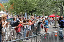 Photographie en couleurs d'une l’athlète entre deux rangées de barrière derrière lesquelles on voit les spectateurs