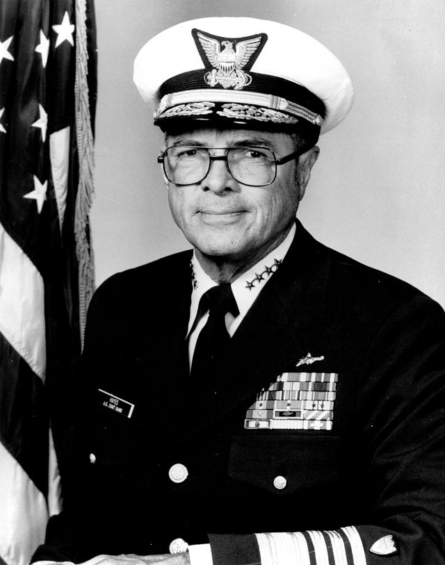 man in Navy uniform with cap, American flag in background