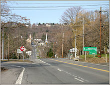 A pair of highways meet at a signalized intersection. In the background at the base of a hill is a small village. One highway leads from the intersection, heads through the village, and ascends a larger hill in the distance.