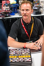 Photographie en couleurs représentant le cinéaste assis, devant une table et des piles de livres intitulés Comic Con.
