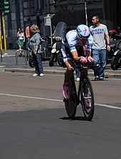 Ein Straßenrennfahrer, der einen hellblauen Skinsuit und einen passenden aerodynamischen Helm mit rosa Schuhen und Handschuhen trägt.  Zuschauer schauen am Straßenrand zu.