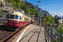 A preserved RAe TEE II trainset passing through  Veytaux-Chillon station, on the Cisalpin's route (but not a stop), with Chillon Castle in the background