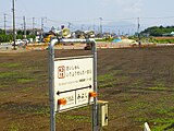 駅名標と再開発中の旧駅跡（左奥）と移転後の御代志駅（右奥）（2023年6月）