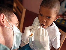 Seorang pria memegang nampan plastik dengan bahan cokelat di dalamnya dan menempelkan tongkat kecil ke mulut yang terbuka dari seorang anak laki-laki