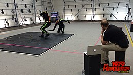 Two actors perform in dark suits on a motion capture stage, surrounded by cameras. The director looks on from the side.