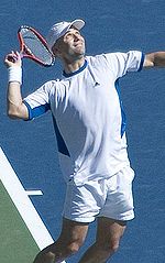 A man in white and blue shorts and T-shirt with a white baseball cap, raises his right arm, holding a modern version of a tennis racket, as he prepares to serve the unseen tennis ball