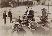 Photographie montrant un homme et une femme élégamment vêtus à bord d'une voiture en stationnement.