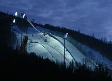 The ski jumping and flying hills at Certak in Harrachov, 2011. The modern curvature of the reprofiled slope can be seen on the flying hill (top), which used to be a much steeper drop. Certakbynight.jpg