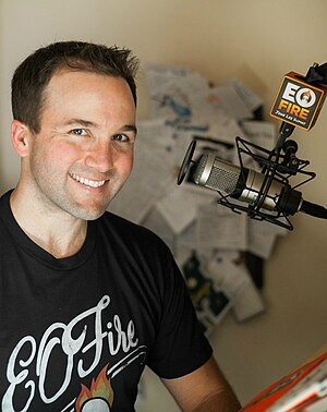 Image of a smiling John Lee Dumas wearing a black t-shirt that says "E O Fire" with a mic seen to the right