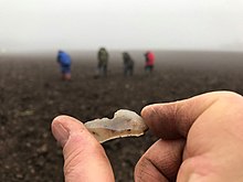 a hand holding a lithic with fieldworkers in the background