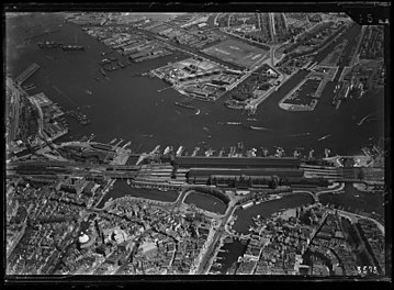 Luchtfoto van het IJ en omgeving, gezien in noordelijke richting. Op de voorgrond het het Damrak en het Stationseiland, op de achtergrond Amsterdam-Noord; circa 1930. Luchtvaartafdeeling, 1920-1940.