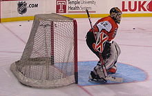 An ice hockey goaltender is on the ice in front of the net, viewed from the right side. He is wearing an orange and white sweater, and black pants.